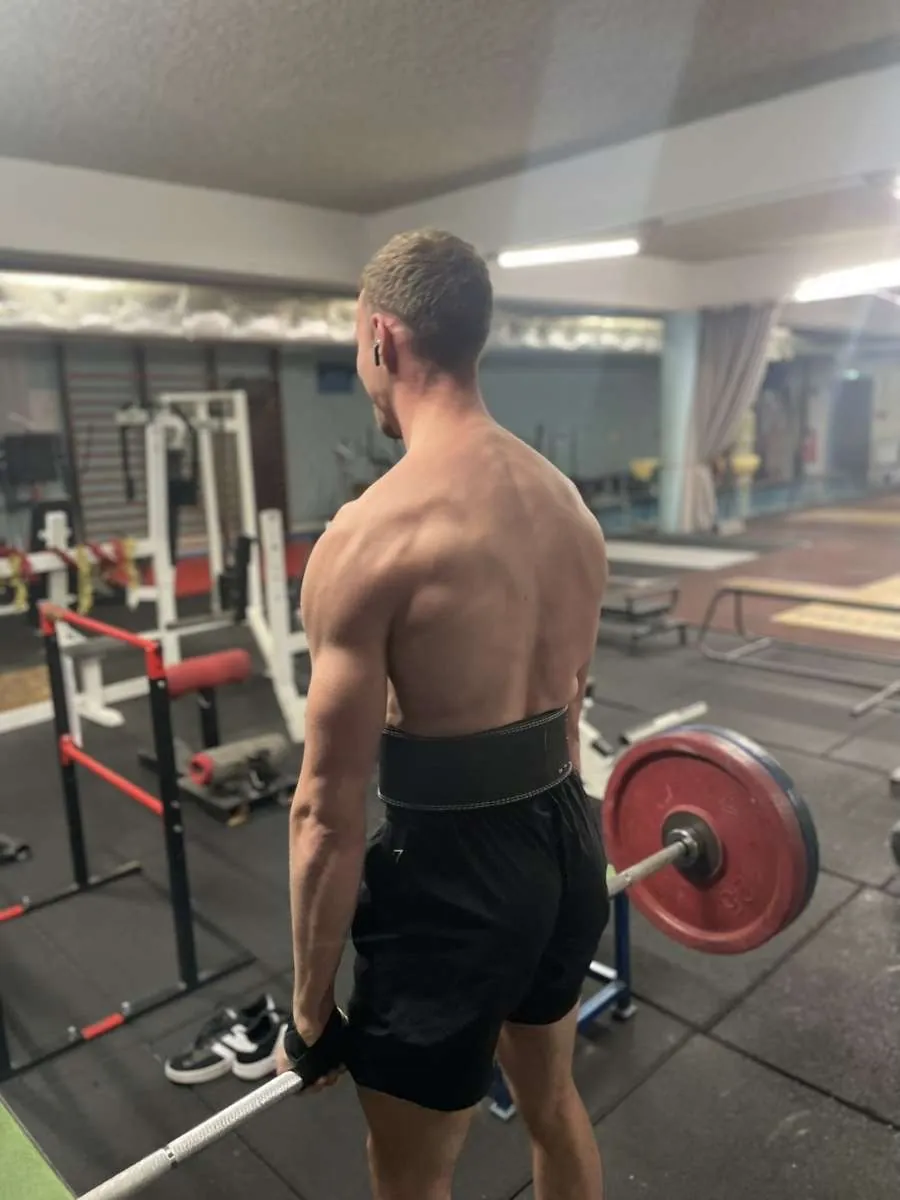 Le coach de dos, devant une barre chargée en salle de musculation